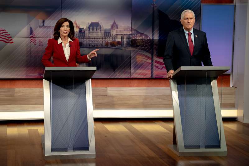 Gov. Kathy Hochul, wearing a red jacket, points toward Bruce Blakeman as both candidates stand at podiums during the Hochul-Blakeman debate in New York.
